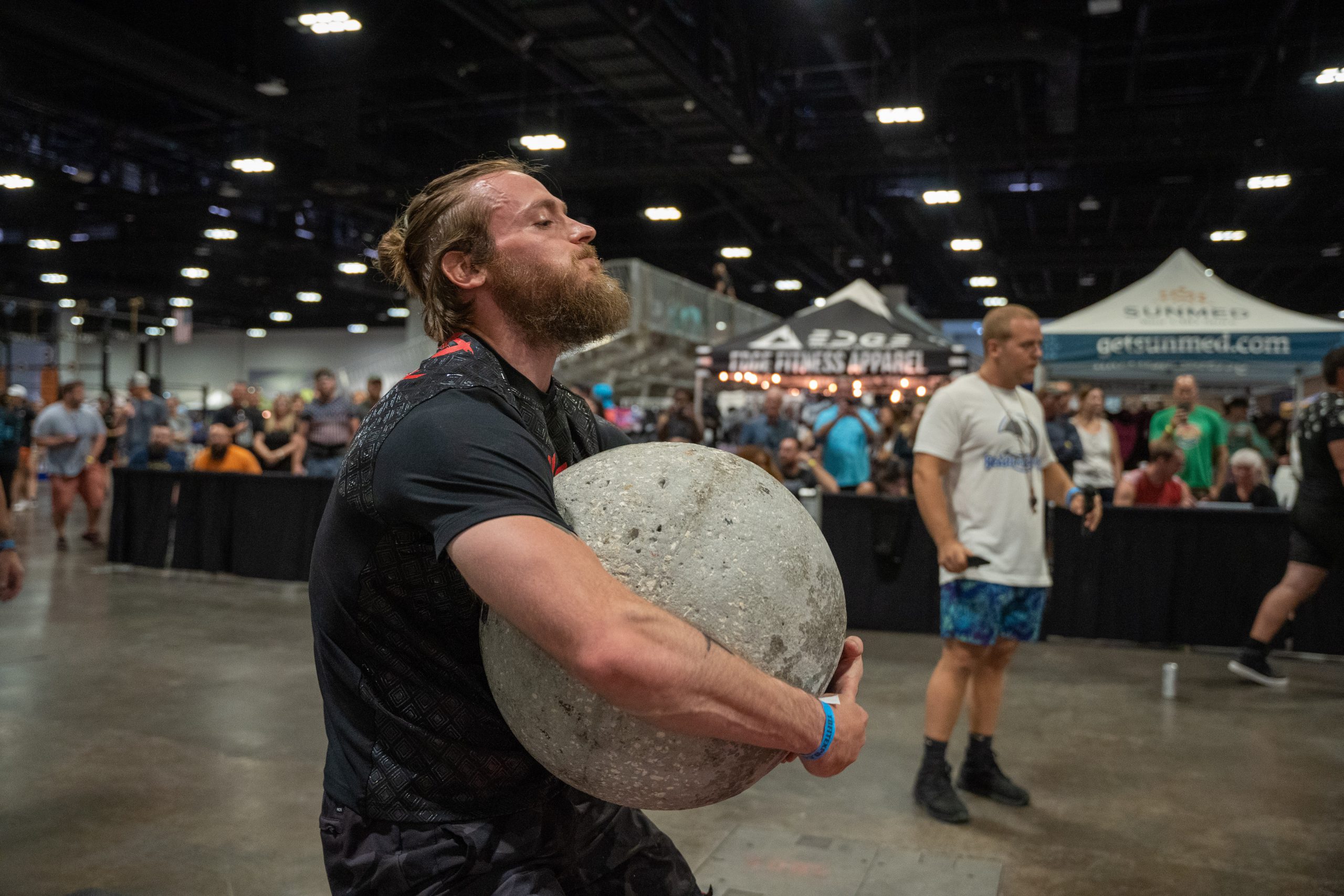 <img src="strongman_atlas_stone.jpg" alt="A strongman athlete lifting an atlas stone onto their shoulder repeatedly for reps. The athlete is shown with a wide stance, squatting down to grip the round stone placed on the ground. They are exerting great strength as they lift the stone off the ground and transition it to their shoulder in a controlled motion. The athlete's muscles are visibly engaged, showcasing their power and determination.">