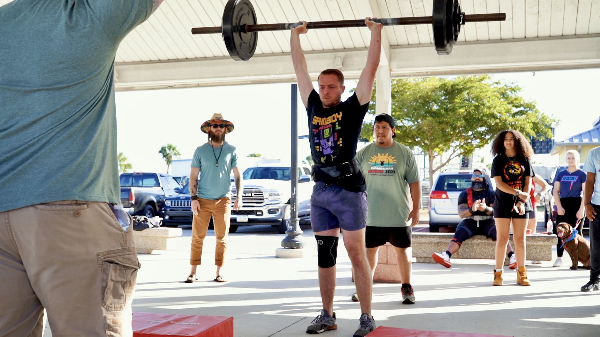 <img src="strongman_split_jerk.jpg" alt="A strongman athlete performing a split jerk lift. He is in a powerful stance, with one leg forward and the other extended back. He is holding a weighted barbell overhead with locked arms, demonstrating strength and stability.">